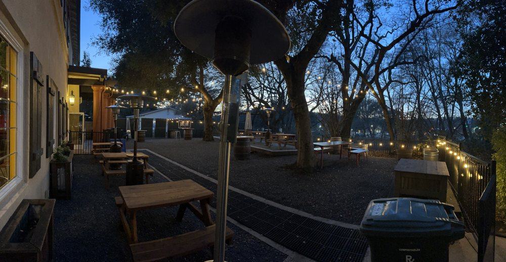 El patio junto al arroyo al anochecer, con luces colgantes entre los robles
